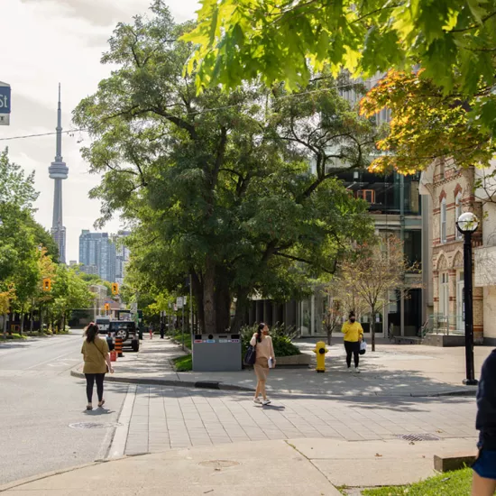 People wearing face masks walking along tree-lined street with the CN Tower in the background. Street sign reads Ursula Franklin St.