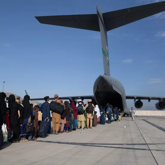 Back of plane on runway with cargo bay door open with a long line of women, men and children, some carrying backpacks or holding suitcases, in civilian clothes heading onto the plane.
