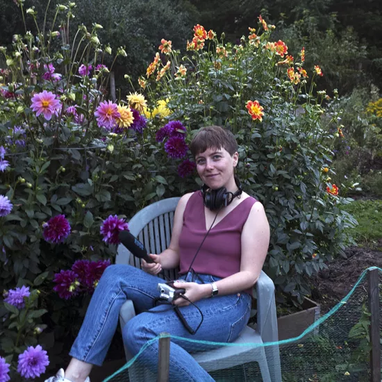 Lauren Ramsay sitting in a patio chair in a garden surrounded by bushes and flowers, wearing jeans and a sleeveless purple shirt, holding a microphone, audio recorder and wearing large headphones around her neck