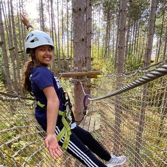 Divya Dey wearing a white helmet standing on a large net strung between two trees. She is hooked up to a cable with a line attached to a belt.