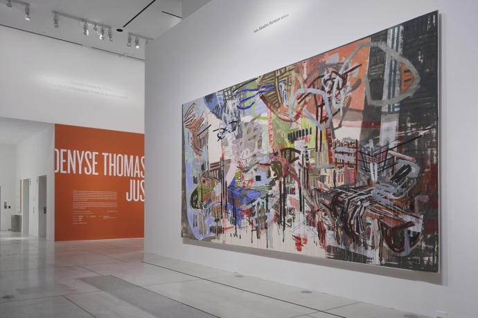 large abstract painting on a gallery wall. In the background on a separate wall is a large sign that reads Denyse Thomasos: Just Beyond