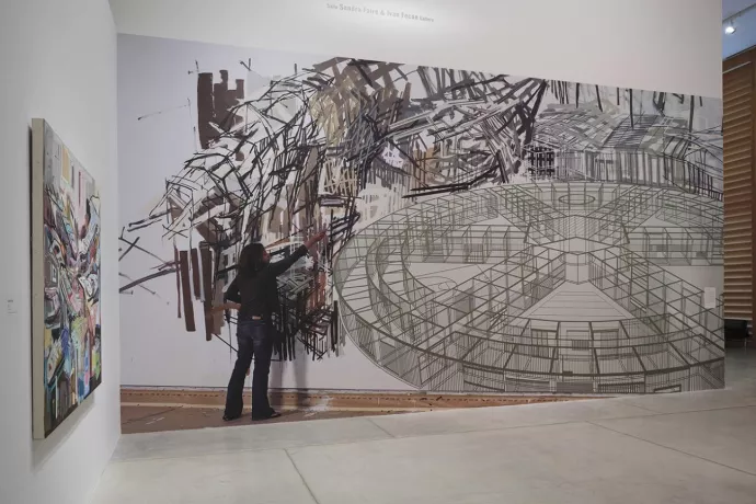large painting on a wall in gallery showing a woman wearing jeans and a black shirt with a paintbrush painting black, abstract lines. The painting includes a large, circular element with spokes moving in toward the centre, resembling a drawing of the inside of a building