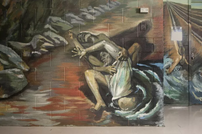 Painting on a cinder block wall depicting a man reaching out from what appears to be water, with another man behind holding him