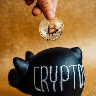 photo of a single bitcoin being put into a black piggy bank that says cryptos on it
