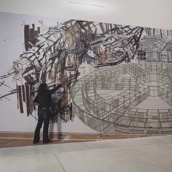 large painting on a wall in gallery showing a woman wearing jeans and a black shirt with a paintbrush painting black, abstract lines. The painting includes a large, circular element with spokes moving in toward the centre, resembling a drawing of the inside of a building
