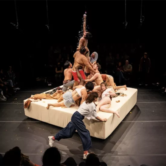Performers on a revolving, raft-like stage. Bodies piled on top of one another