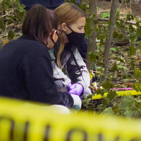 two students crouched on the ground in the woods with yellow caution tape in the foreground