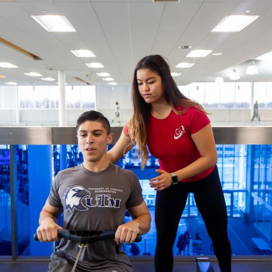 A male student on a rowing machine with a student personal trainer supporting them