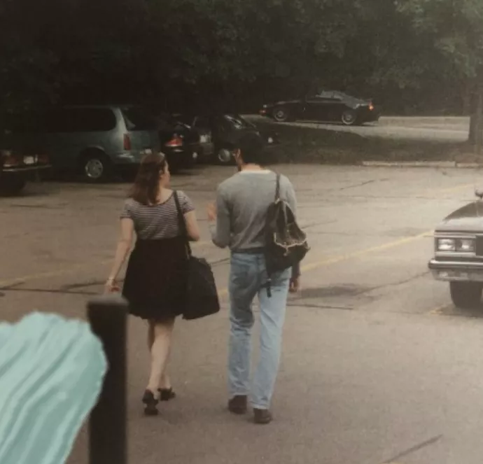 A couple walks in a parking lot with a 1970s car parked off to the side
