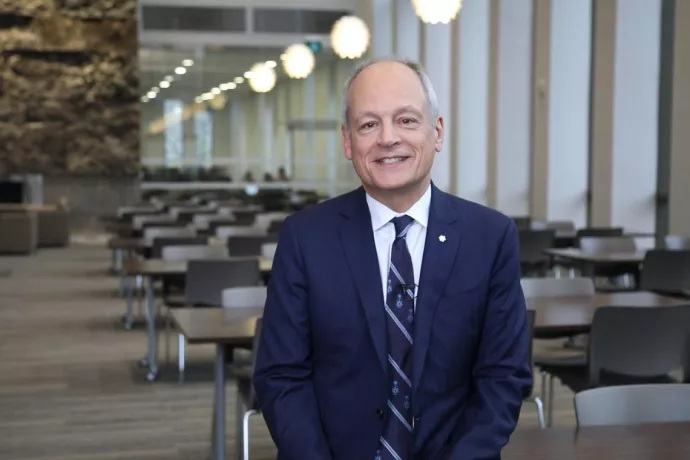 Image of Meric Gertler in a blue suit standing in a long room with tables and a bank of windows to one side