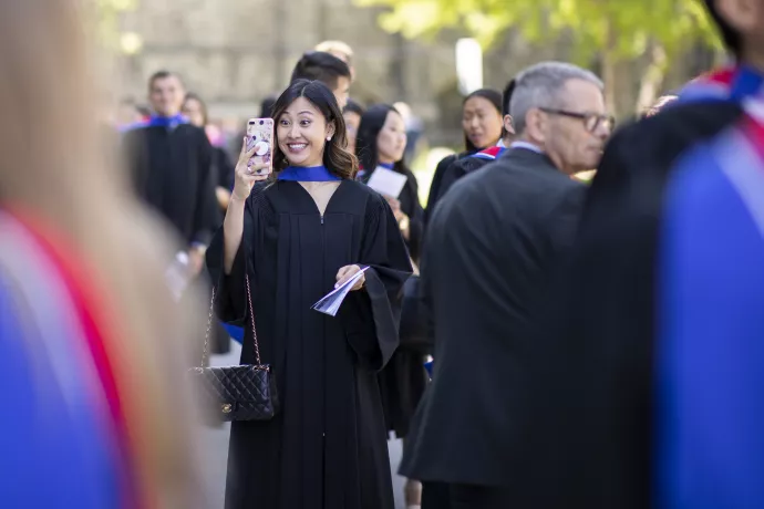 A woman in convocation regalia smiles as she takes a selfie photo with her phone.