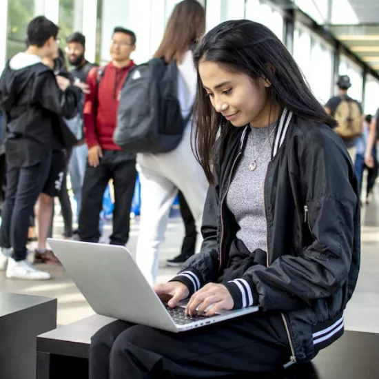 Female student in UTM's CCT building on her laptop with other students in the background