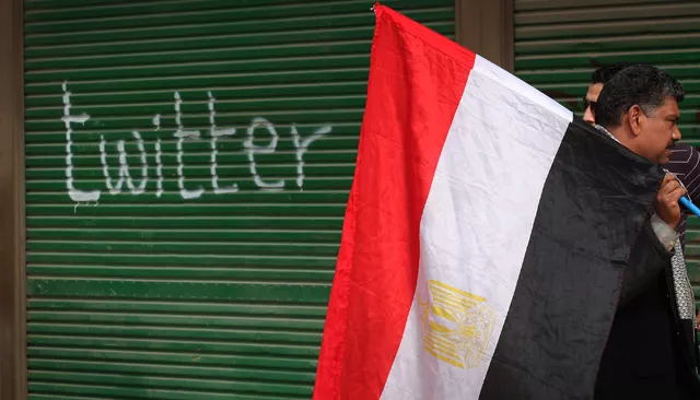 Photograph of middle-aged man carrying Egyptian flag past a garage on with the word twitter has been spraypainted