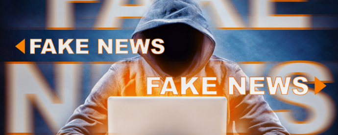 A hooded figure, face hidden, is seated in front of a laptop.  The words "fake news" are are in white above the image of the person.