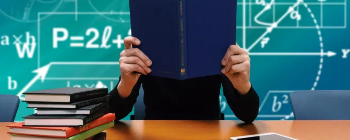 A person is holding up a book in front of their face. There are books on the table in front of them and a blackboard with a variety of symbols on it behind them.