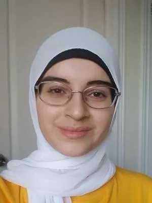 A woman smiling, wearing a white hijab over a yellow shirt and metal-framed glasses.