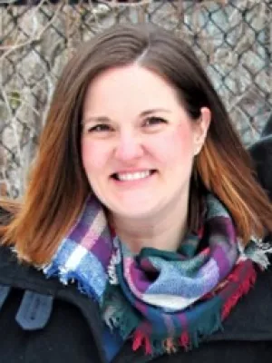 A woman smiling, with medium-length brown hair, wearing a plaid scarf over a black winter jacket.