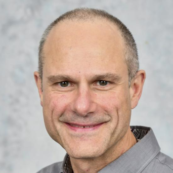 A man smiling, with short, grey hair, wearing a grey shirt.