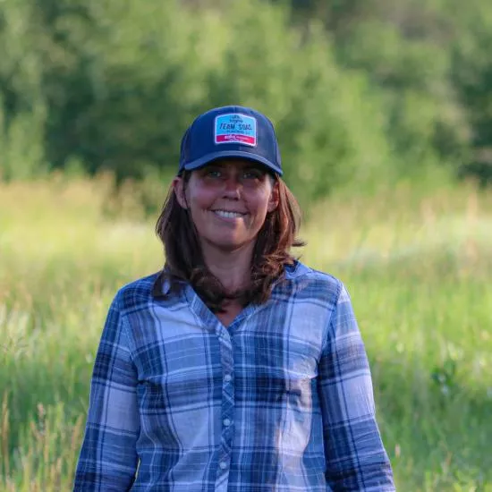 A woman smiling, standing in a field of tall grass, with medium-length brown hair, a blue baseball cap, and a blue flannel.