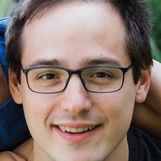 A man smiling, with short, brown hair, wearing black-framed rectangular glasses.