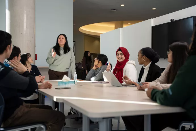 A owman holds a conversation with a group of diverse students at a long table