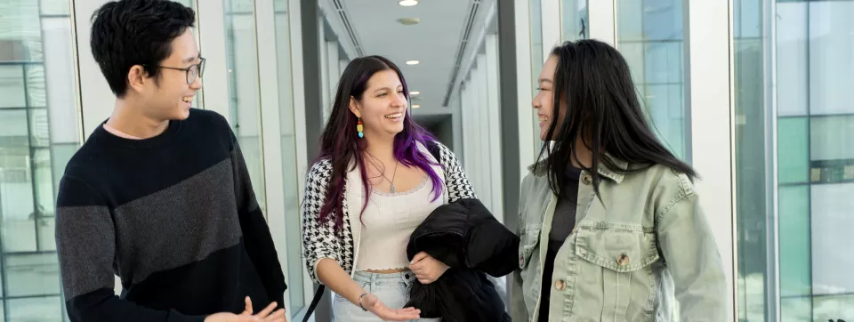 Three students have an animated friendly conversation in a bright hallway