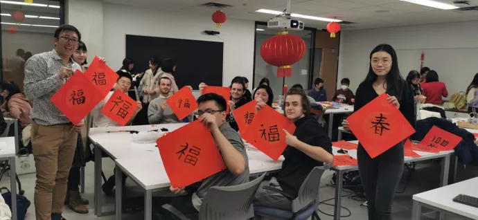 Students and instructors seated around a table and standing beside the table hold up red sheets of paper with Chinese calligraphy