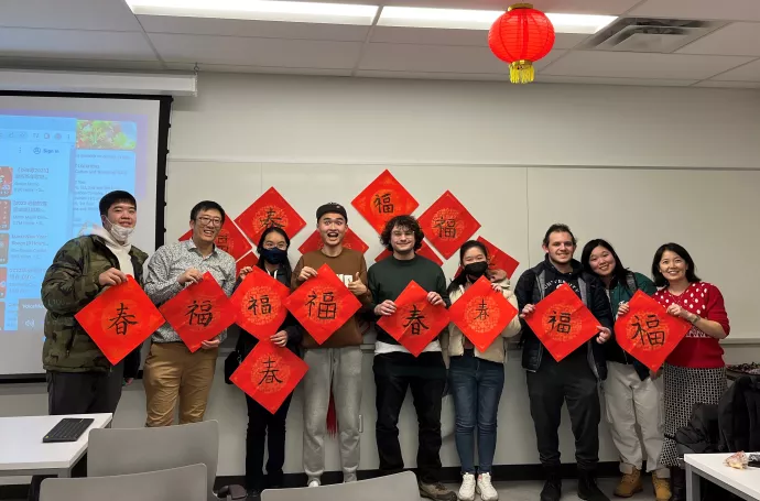 Participants and instructors are standing at the front of a classroom, holding red sheets of paper with the Chinese characters for "spring" or "luck" written in calligraphy