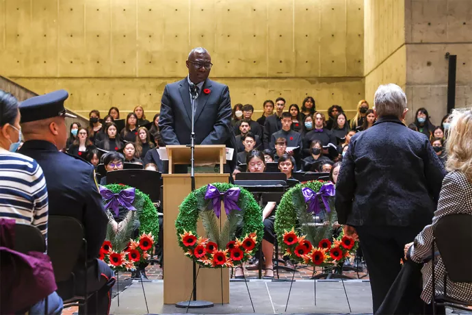 Man stands at podium with poppy on his jacket, choir stands behind him