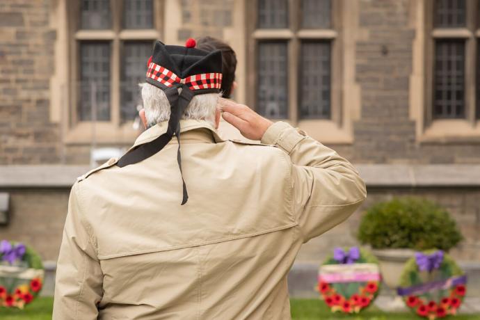 Man standing with back to camera saluting row of poppy wreaths on ground in front of him.