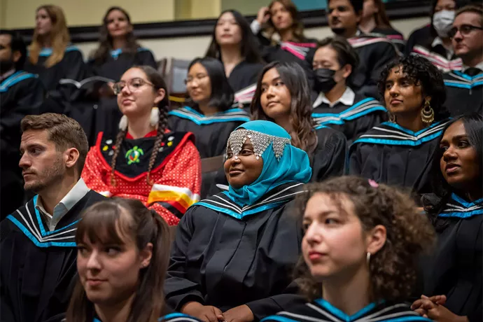 Graduates wearing black gowns and light blue stoles sitting in Convocation Hall.