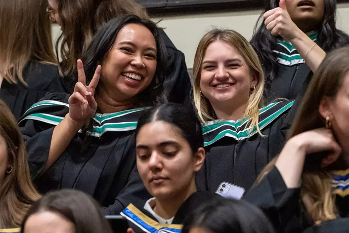 Close up of two graduates sitting in Convocation Hall, both smiling at the camera, one holding up their hand giving the peace sign.