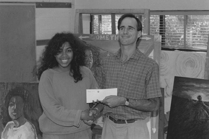 Black and white photo of Denyse Thomasos and John Armstrong holding a white piece of paper between them as they smile for the camera. Artwork stacked against a wall can be see in the background.