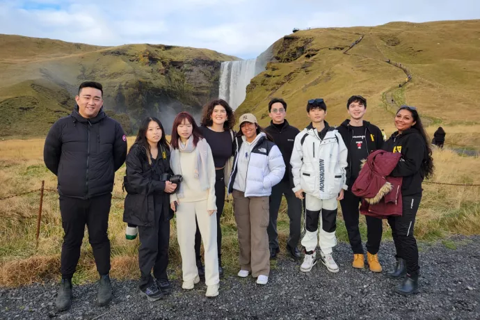 A group of nine students stand in a row in front of a yellow-green hill and tall, narrow waterfall.