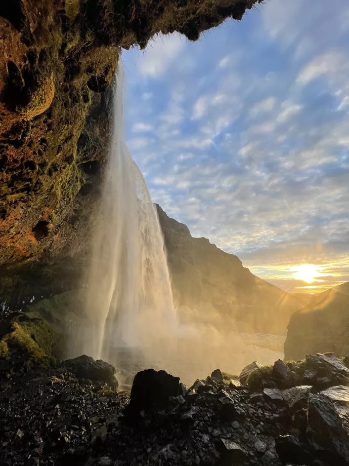 Waterfall from below, flowing down from a curving rock face with jagged rocks along the bottom and the setting sun in the background