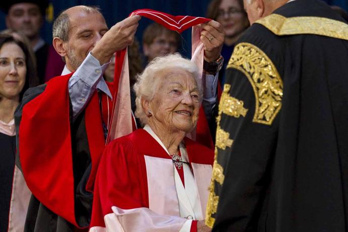 Professor Ulli Krull stands behind Hazel McCallion, draping a red hood over her shoulders
