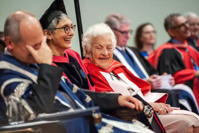 Hazel McCallion sits in a row with others, looking over in the direction of the camera as she laughs. Two seats from her is President Meric Gertler laughing with his hand in front of his face.