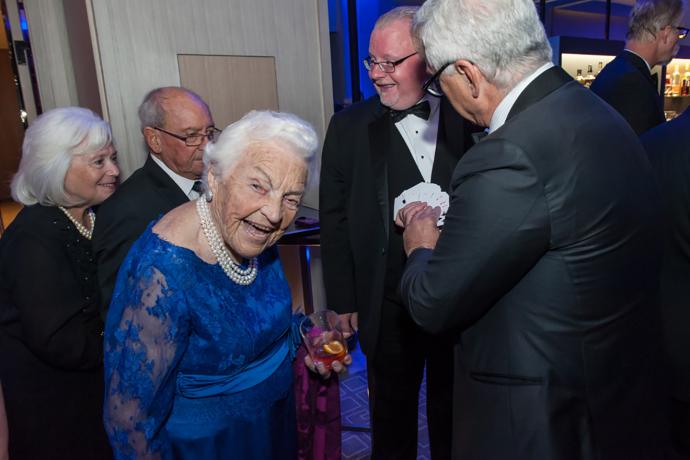 Hazel McCallion stands with a small group of people at a gala, looking at the camera with a big smile on her face as a man next her her holds out a deck of cards