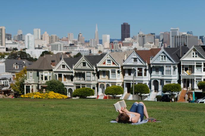 Woman laying in grass reading a book in a park, in background is row of colourful houses and farther back are taller buildings