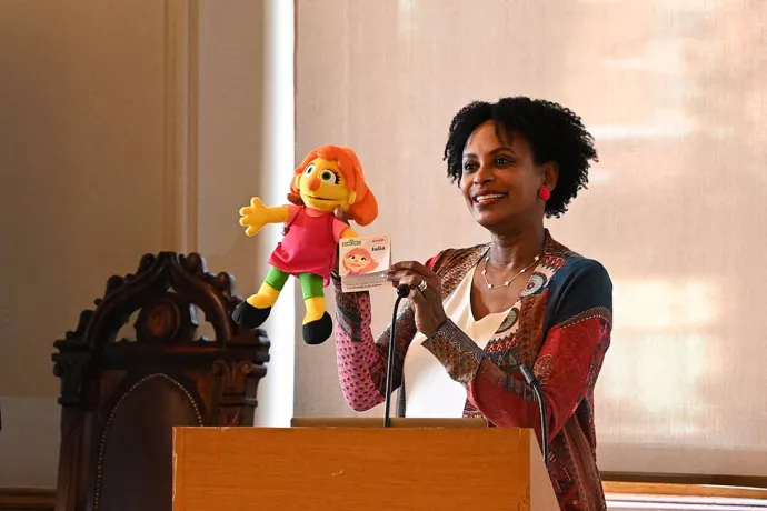 Rhonda McEwen stands at a podium holding up a muppet with orange hair, a pink top, green pants and black shoes
