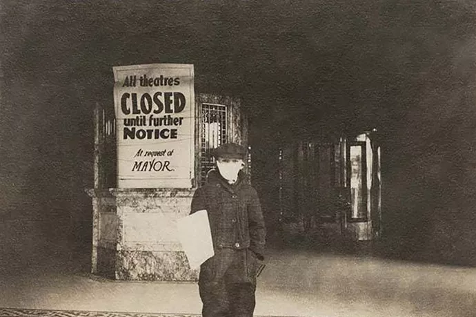 Black and white photo of a newspaper boy wearing a cloth face mask standing outside a theatre with a large sign over the ticket booth that reads "All theatres closed until further notice at request of mayor" 