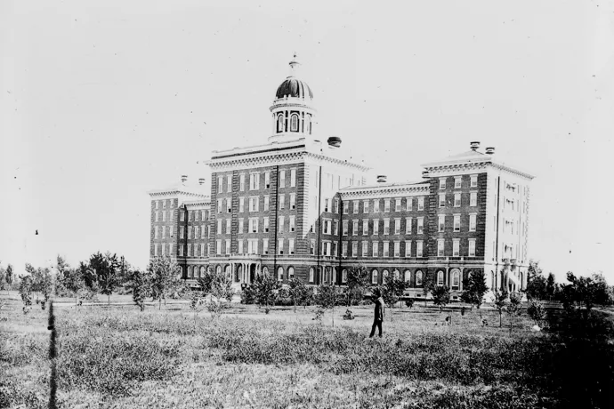 Black and white photo of a large brick building in a field with three wings. 