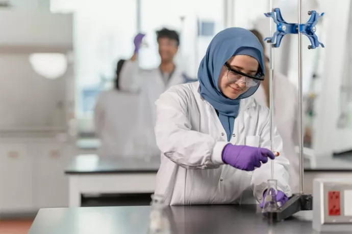 A woman in a lab coat wearing protective goggles measures a solution in a beaker