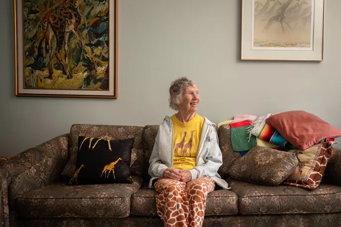 Anne Innis Dagg sitting on a couch wearing giraffe printed pants with portaits of giraffes on the wall behind her.