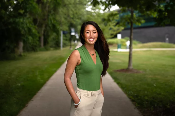 Woman with long dark brown hair wearing a light green shirt and white pants standing on a tree-lined pathway looking directly at the camera 