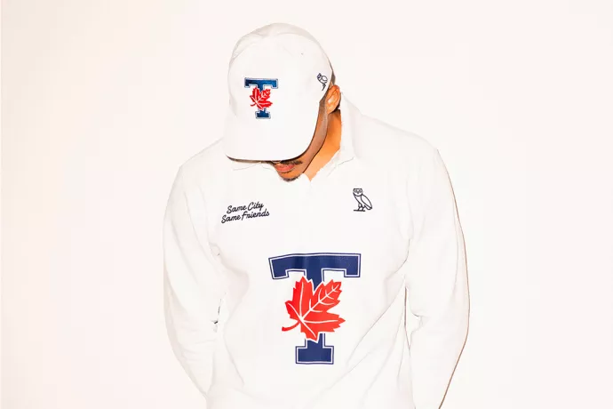 Man wearing white ball cap with blue T overlaid with red maple leaf with line drawing of owl on the side. Also wearing a hood with same logo, blue T overlaid with red maple leaf, line drawn owl on upper right chest, written in cursive on left is: Same city, same friends