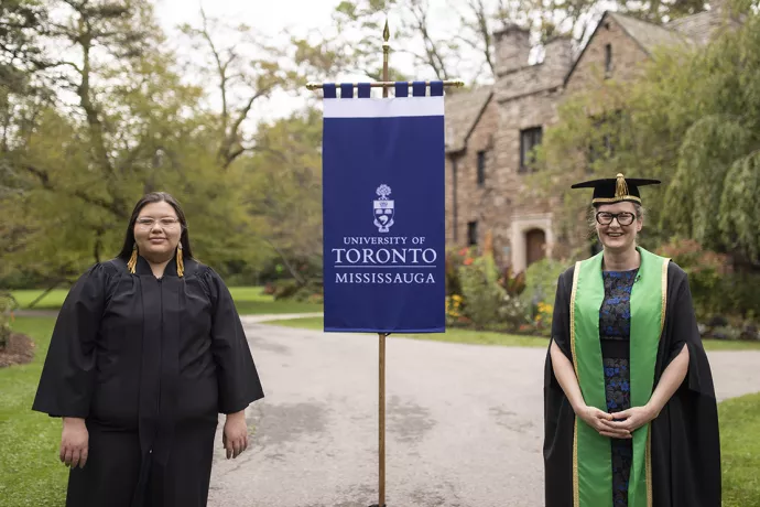 Kaeliana Smoke wearing a black robe and and Alexandra Gillespie wearing a black robe and green sash stand with a blue flag between them that reads University of Toronto Mississauga