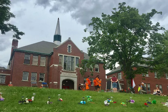 Stuffed toys laid out on lawn in front of Kamloops Indian Residential School, people standing in front of building wearing orange t-shirts