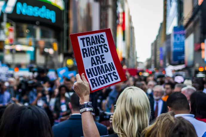 Protestors fill street, with tall buildings lining either side of the street. In foreground is a person holding up a sign that reads: Trans rights are human rights