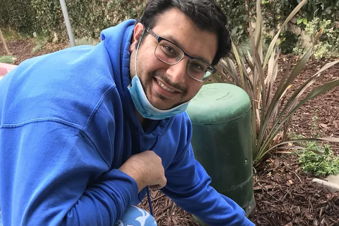 Jamsheed wearing a blue hoodie, glasses and blue face mask pulled down, smiling at camera while kneeling on a pathway next to greenspace with mulch on the ground.. 